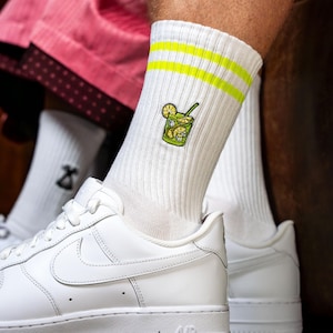 Könnte beinhalten: Weiße Socken mit gelben Streifen und einem gestickten Bild eines Glases Limonade. Die Socken werden an den Füßen einer Person mit weißen Turnschuhen getragen.