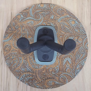Top-down view of a circular wooden guitar hanger base featuring a laser-engraved teal and tan paisley pattern with a black padded wall mount yoke in the center.