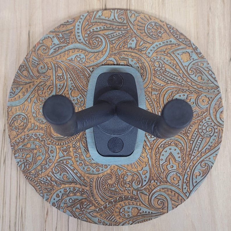 Top-down view of a circular wooden guitar hanger base featuring a laser-engraved teal and tan paisley pattern with a black padded wall mount yoke in the center.