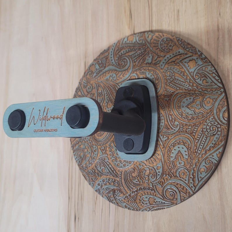 Angled side view of a custom guitar wall mount showing the depth of the black padded hooks and the intricate textured engraving on the circular wooden backplate.