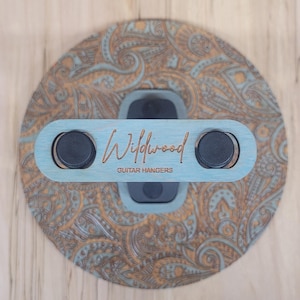 Top-down view of a circular wooden guitar hanger base featuring a laser-engraved teal and tan paisley pattern with a black padded wall mount yoke in the center.