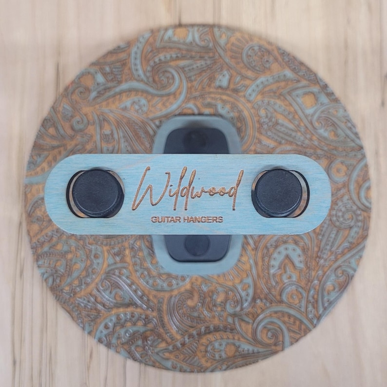 Top-down view of a circular wooden guitar hanger base featuring a laser-engraved teal and tan paisley pattern with a black padded wall mount yoke in the center.