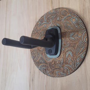 Angled side view of a custom guitar wall mount showing the depth of the black padded hooks and the intricate textured engraving on the circular wooden backplate.