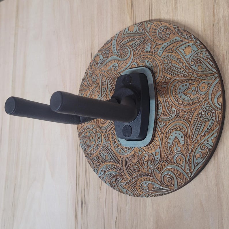 Angled side view of a custom guitar wall mount showing the depth of the black padded hooks and the intricate textured engraving on the circular wooden backplate.