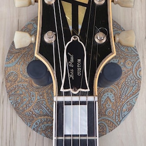 Top-down view of a guitar hanging on a circular wooden guitar hanger base featuring a laser-engraved teal and tan paisley pattern with a black padded wall mount yoke in the center.
