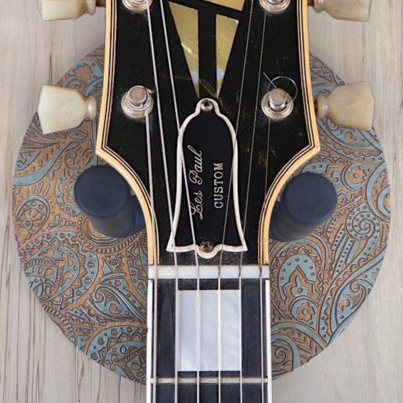 Top-down view of a guitar hanging on a circular wooden guitar hanger base featuring a laser-engraved teal and tan paisley pattern with a black padded wall mount yoke in the center.