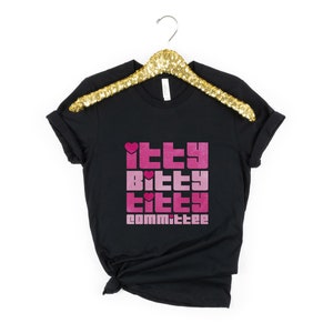 May include: Black t-shirt with the text "Itty Bitty Titty Committee" in pink and magenta block letters. The shirt is hanging on a gold sequined hanger against a white background. The text includes heart symbols.