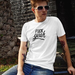 May include: White t-shirt with the text "FUCK YEAH" in black script. The shirt is paired with blue jeans. The person is wearing sunglasses.