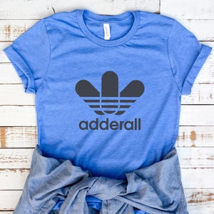 May include: A light blue t-shirt with a black graphic design resembling a stylized flower and the word "Adderall" in a sans-serif font. A denim shirt is draped below the t-shirt on a white wooden surface.