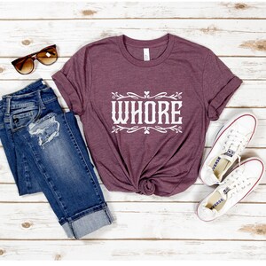 May include: A maroon t-shirt with the word "WHORE" in white, stylized text. The shirt is paired with blue denim jeans, white sneakers, and brown sunglasses. The jeans have distressed details.