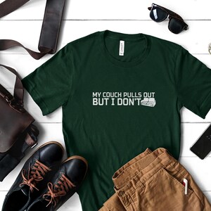 May include: Dark green t-shirt with the text "MY COUCH PULLS OUT BUT I DON'T" in white. The shirt is displayed with brown pants, black shoes, a leather bag, sunglasses, and a smartphone on a white wooden surface.