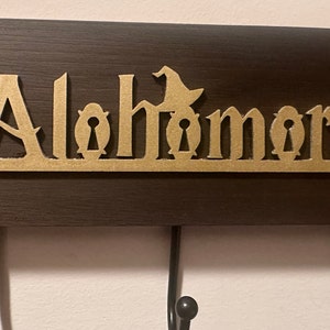May include: A wooden key holder with the word "Alohomora" carved in gold lettering. The word is surrounded by keyhole designs and other magical symbols. The holder has three hooks for hanging keys.