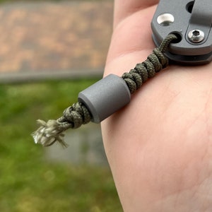 Può includere: Un coltellino tascabile grigio con un laccio nero e una perla grigia. Il coltello è chiuso e ha una lama parzialmente visibile.