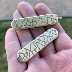 Op de afbeelding: Twee messing zakgereedschappen met een steenachtig patroon worden in een hand gehouden. De gereedschappen zijn rechthoekig met afgeronde uiteinden en een kleine bevestiging. Het oppervlak van de gereedschappen heeft een getextureerd, onregelmatig ontwerp.