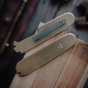 May include: Two brass tools with a black handle. The top tool has a small pry bar and a rounded end. The bottom tool has a rounded end and a cross etched into the surface. The tools are on a wooden surface.