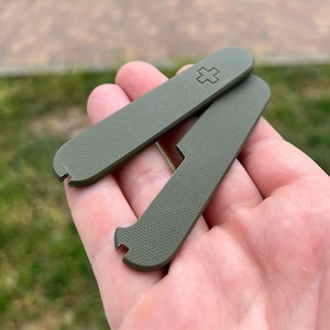 May include: Two green plastic scales for a Swiss Army knife. The scales have a textured surface and a small cross embossed on one of the scales.