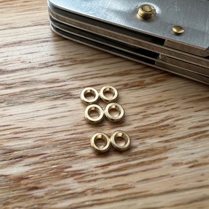 Può includere: Primo piano di sei piccoli anelli di metallo dorati su una superficie di legno. Gli anelli sono disposti in due file di tre.