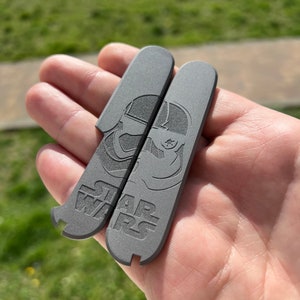 Könnte beinhalten: Zwei graue Metall-Schlüsselanhänger mit einem Star Wars Stormtrooper-Design und dem Text "STAR WARS" darauf.