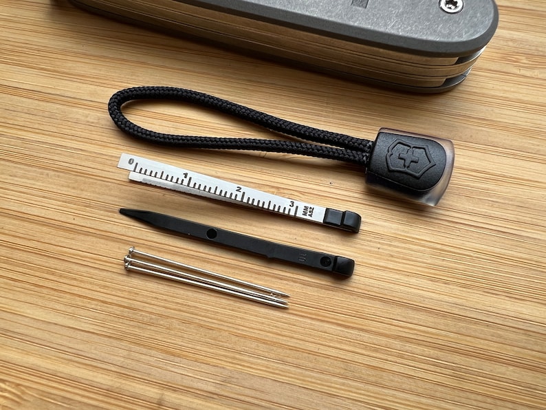 Pode incluir: Um conjunto de ferramentas para uma faca multiferramentas, incluindo um cord&atilde;o preto com o logotipo do ex&eacute;rcito su&iacute;&ccedil;o, uma pequena r&eacute;gua marcada em cent&iacute;metros, um furador preto e tr&ecirc;s agulhas de costura prateadas.