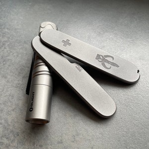May include: A silver pocket knife with a cross and a Mandalorian symbol, along with a silver Olight flashlight. The knife has a small blade and a bottle opener. The items are on a gray surface.