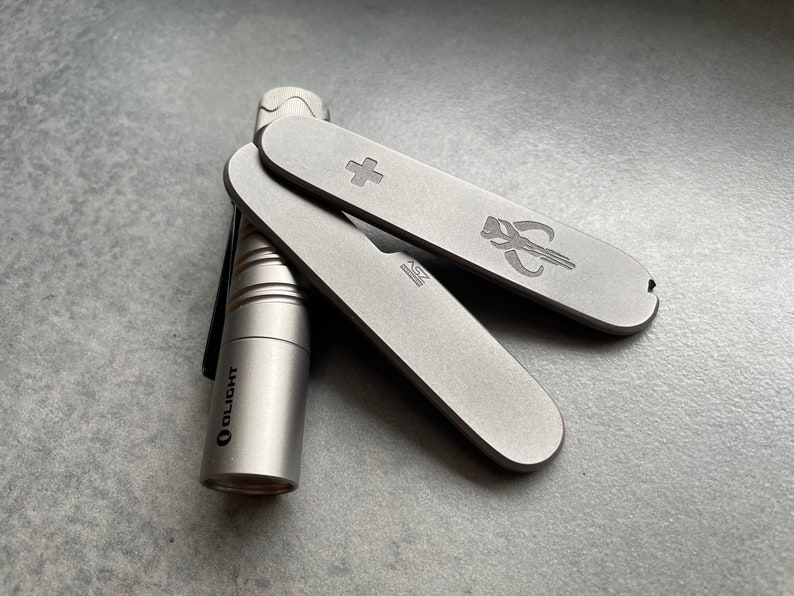 May include: A silver pocket knife with a cross and a Mandalorian symbol, along with a silver Olight flashlight. The knife has a small blade and a bottle opener. The items are on a gray surface.