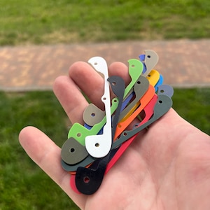 Op de afbeelding: Een verzameling van 10 verschillende gekleurde kunststof vervangende onderdelen voor een mes. De onderdelen zijn in verschillende tinten groen, blauw, rood, oranje, geel en zwart. De onderdelen zijn in een waaier vorm in de hand van een persoon gerangschikt.