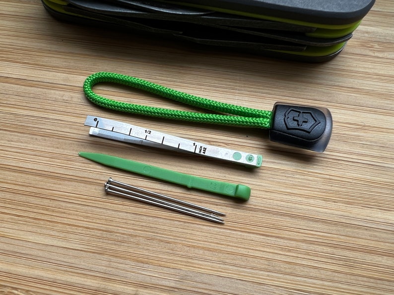 Pode incluir: Um conjunto de ferramentas que inclui um cord&atilde;o verde com um logotipo preto de faca su&iacute;&ccedil;a, uma r&eacute;gua pequena, uma ferramenta de pl&aacute;stico verde e tr&ecirc;s pinos prateados.