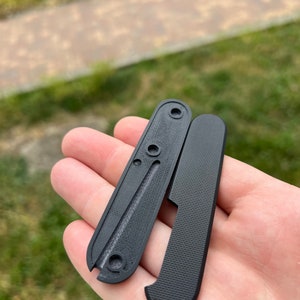 May include: Two black plastic scales for a pocket knife. The scales have a textured surface and are designed to fit a specific knife model.