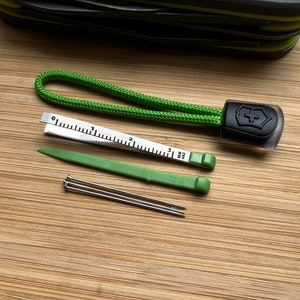 Pode incluir: Um cord&atilde;o verde com um logotipo preto de canivete su&iacute;&ccedil;o, uma pequena r&eacute;gua, uma ferramenta de pl&aacute;stico verde e tr&ecirc;s agulhas de costura.