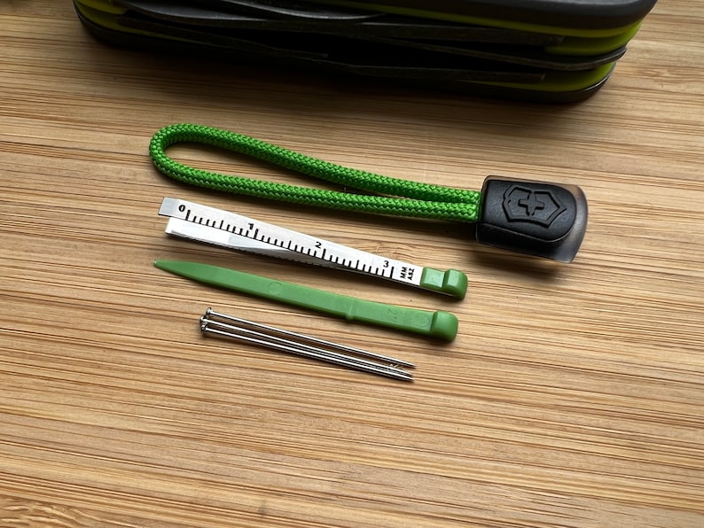 Pode incluir: Um cord&atilde;o verde com um logotipo preto de canivete su&iacute;&ccedil;o, uma pequena r&eacute;gua, uma ferramenta de pl&aacute;stico verde e tr&ecirc;s agulhas de costura.