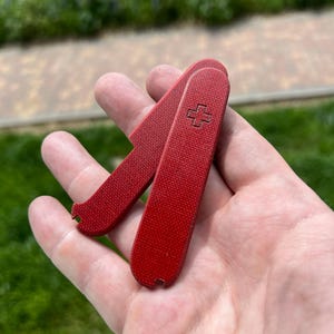 Puede incluir: Una navaja de bolsillo roja con una superficie texturizada, con un emblema de cruz. La navaja está abierta, revelando la hoja. El fondo está borroso, con hierba verde y una zona pavimentada visible.