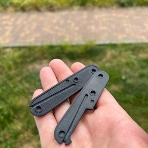 May include: Two black plastic knife scales with a textured surface and multiple holes. The scales are held in a hand.