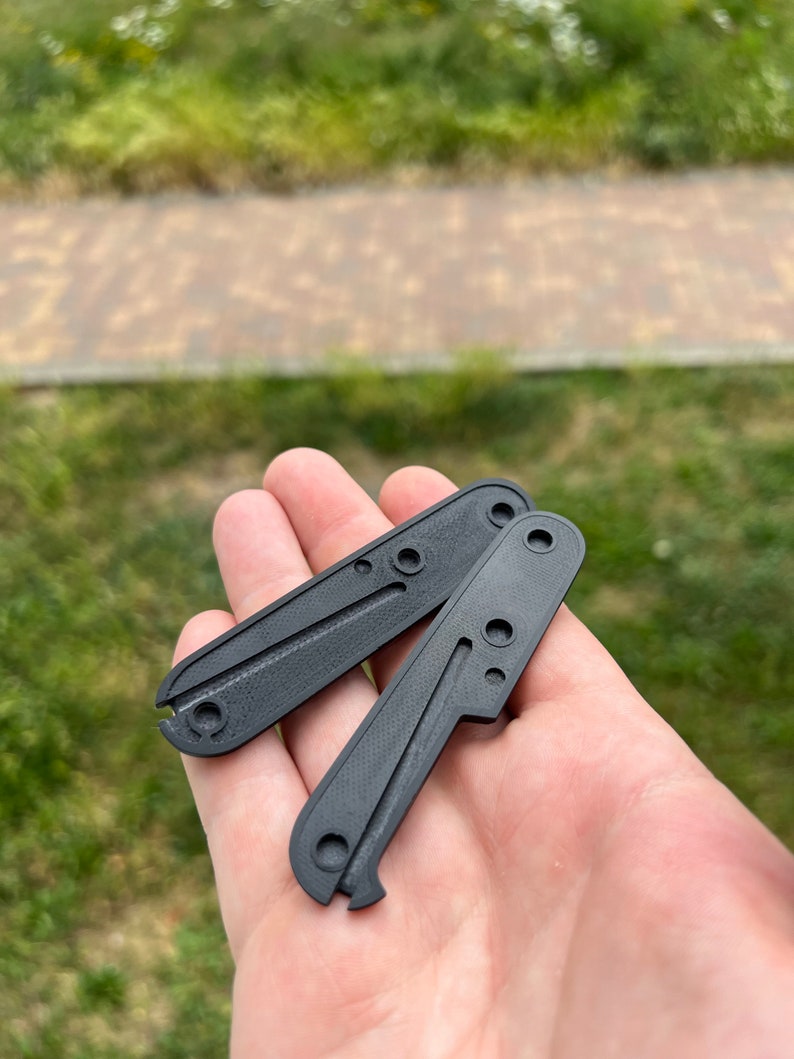 May include: Two black plastic knife scales with a textured surface and multiple holes. The scales are held in a hand.