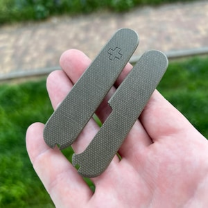 Puede incluir: Dos cachas de navaja de bolsillo verde oliva con una superficie texturizada se sostienen en una mano. Una cacha tiene un símbolo de cruz. Las cachas están diseñadas para reemplazar las cachas originales de una navaja de bolsillo.
