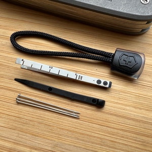 Pode incluir: Um conjunto de ferramentas para um canivete su&iacute;&ccedil;o, incluindo um cord&atilde;o preto com o logotipo do canivete su&iacute;&ccedil;o, uma r&eacute;gua com marcas em polegadas, uma ferramenta preta com um pequeno furo e tr&ecirc;s pinos prateados.