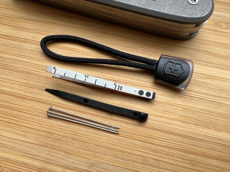 Pode incluir: Um conjunto de ferramentas para um canivete su&iacute;&ccedil;o, incluindo um cord&atilde;o preto com o logotipo do canivete su&iacute;&ccedil;o, uma r&eacute;gua com marcas em polegadas, uma ferramenta preta com um pequeno furo e tr&ecirc;s pinos prateados.