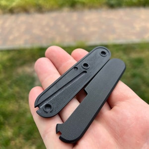 May include: Two black plastic scales for a pocket knife. The scales have a textured surface and a hole for a screw.