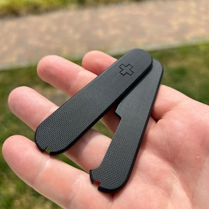 May include: Two black plastic scales for a Swiss Army knife. The scales have a textured surface and a small cross embossed on one of the scales.