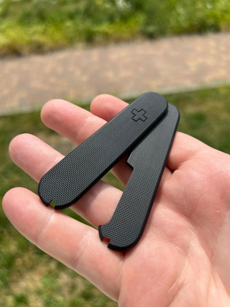 May include: Two black plastic scales for a Swiss Army knife. The scales have a textured surface and a small cross embossed on one of the scales.