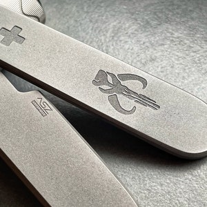 May include: Close-up of a multi-tool with a matte gray finish. The tool features a cross symbol and a Mandalorian emblem. The text "ASZ" and "HANDMADE" are also visible. The tool is designed for everyday use.
