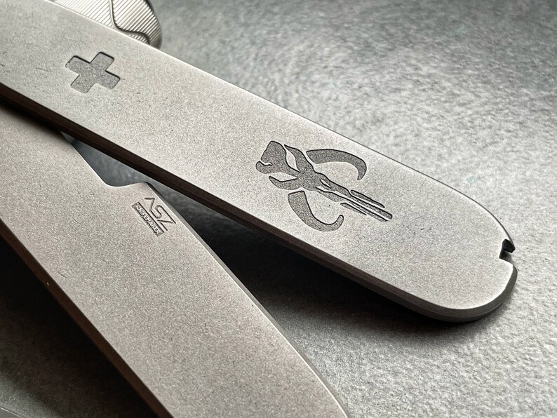 May include: Close-up of a multi-tool with a matte gray finish. The tool features a cross symbol and a Mandalorian emblem. The text "ASZ" and "HANDMADE" are also visible. The tool is designed for everyday use.