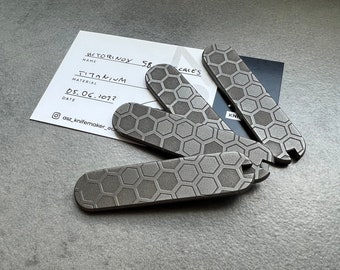 Könnte beinhalten: Vier Titan-Schalen für ein Messer, jede mit einem sechseckigen Muster. Die Schalen sind grau und haben eine gebürstete Oberfläche. Die Schalen sind mit "METORINOX 58" und "CACES" beschriftet.