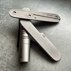 May include: A close-up of a silver-colored multi-tool and a small cylindrical flashlight. The multi-tool has a blade and a flat, rectangular body with several holes. The flashlight is labeled "OLIGHT".