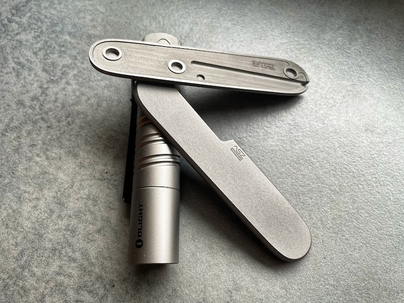 May include: A close-up of a silver-colored multi-tool and a small cylindrical flashlight. The multi-tool has a blade and a flat, rectangular body with several holes. The flashlight is labeled "OLIGHT".