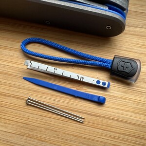 Pode incluir: Um conjunto de ferramentas que inclui uma corda azul com um logotipo Victorinox preto, uma ferramenta de pl&aacute;stico azul, uma r&eacute;gua com marcas em polegadas e cent&iacute;metros, e tr&ecirc;s pinos prateados.