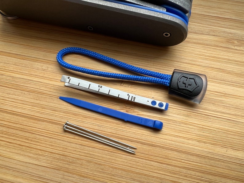 Pode incluir: Um conjunto de ferramentas que inclui uma corda azul com um logotipo Victorinox preto, uma ferramenta de pl&aacute;stico azul, uma r&eacute;gua com marcas em polegadas e cent&iacute;metros, e tr&ecirc;s pinos prateados.