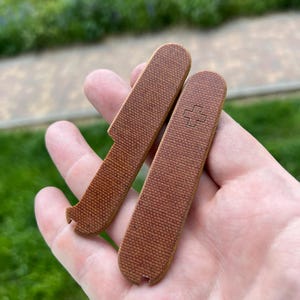 Puede incluir: Dos cachas de cuchillo marrones con textura de lona. Una cacha tiene una muesca y un extremo curvado. La otra cacha tiene un símbolo de cruz. Las cachas se sostienen en una mano sobre un fondo verde borroso.