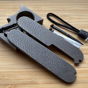 Könnte beinhalten: Ein Multitool mit einer strukturierten, grauen Metalloberfläche. Das Werkzeug verfügt über einen Flaschenöffner, einen Schlitzschraubendreher und einen Kreuzschlitzschraubendreher. Es enthält auch eine kleine, schwarze Schnur mit einem Metalletikett.