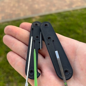 May include: A black multi-tool with a silver blade and a green tool. The tool is open and the blade is extended. The tool is held in a hand.