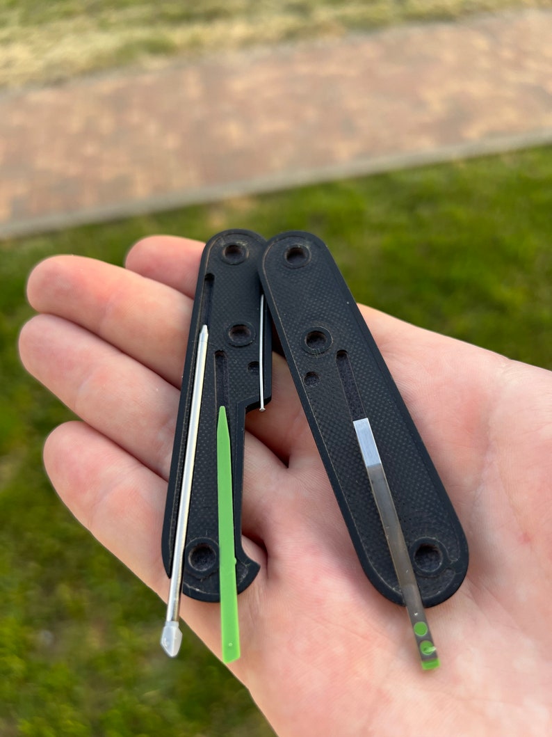 May include: A black multi-tool with a silver blade and a green tool. The tool is open and the blade is extended. The tool is held in a hand.